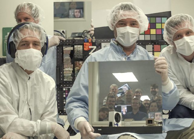 A photo taken by one of the flight cameras shows, left to right: Andy Winhold, Justin Maki, Jim Bell and Alex Hayes. Bell is holding a mirror reflecting the faces of additional team members looking into the cleanroom window from the data analysis room outside.